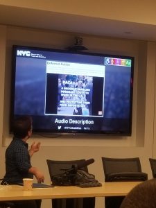 Jonathan, a little person stands in front of a tv screen with a slide describing how to make social media accessible using ALT Text.
